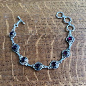 May include: A silver-toned bracelet featuring six circular settings, each containing a deep red cabochon stone. The links are connected by small metal rings, and the piece includes a toggle clasp closure at one end and three adjustment rings.