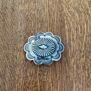 May include: A silver-toned concho with a geometric design. The concho has a scalloped edge and a raised center.