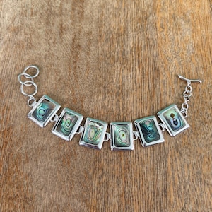 May include: Silver-toned bracelet featuring rectangular abalone shell inlays. The iridescent shells are set in a silver frame and connected by small links. The bracelet has a toggle clasp. The jewelry is displayed on a wooden surface.