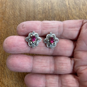 May include: A pair of silver earrings with a pink gemstone in the center of each earring. The earrings are in the shape of a flower with a detailed design.