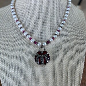 May include: A pearl necklace with a red and silver pendant. The necklace features white pearls interspersed with small red beads. The pendant is a small, round purse design with red and silver accents. The necklace is displayed on a neutral-colored surface.