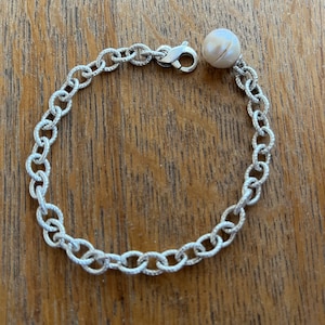May include: A silver chain bracelet with a single pearl charm. The pearl is off-white and has a slight iridescent sheen.