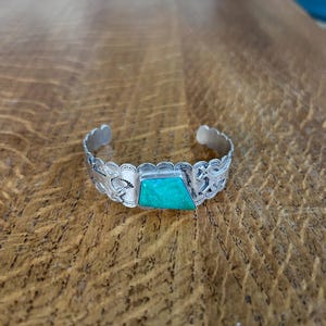 May include: A silver-toned cuff bracelet with a turquoise-colored stone. The bracelet has a decorative pattern along the edges and around the stone. The stone is a trapezoid shape. The bracelet is displayed on a wooden surface.
