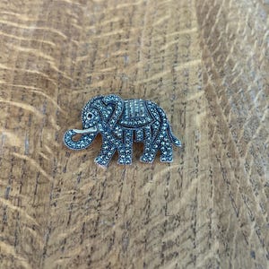 May include: A silver-toned elephant brooch with intricate detailing. The brooch features a textured surface with small, sparkling embellishments. The elephant has a raised trunk and a decorative saddle. The brooch is a small, detailed piece of jewelry.