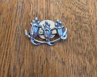 May include: Silver tone flower brooch with three stylized flowers. The flowers are arranged in a row with a stem connecting them.