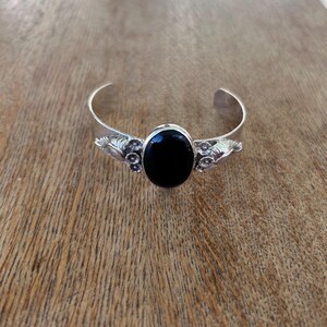 May include: A silver cuff bracelet with an oval, black gemstone center. The bracelet has decorative floral and leaf accents on each side of the gemstone. The bracelet is displayed on a wooden surface.