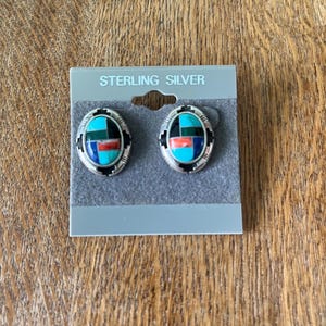 May include: Oval-shaped sterling silver earrings with inlaid turquoise, red, and blue stones. The earrings are on a gray card that reads "STERLING SILVER". The earrings have a black border and are displayed on a wooden surface.