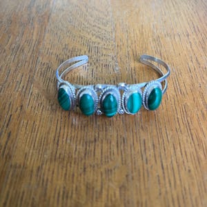 May include: A silver cuff bracelet featuring five oval-shaped, vibrant green malachite stones. The stones are set in individual bezels and connected by a delicate silver band. The bracelet is displayed on a wooden surface.