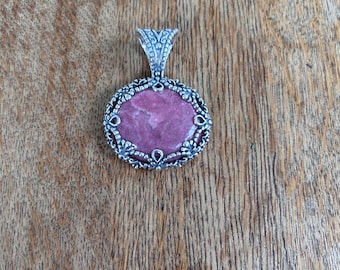 May include: A silver-toned pendant with an oval, pink gemstone. The gemstone is set in a decorative, openwork frame. The pendant has a detailed bail for attaching to a chain. The background is a wooden surface.