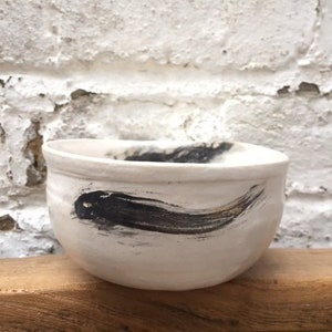 May include: White ceramic bowl with a black and gold brushstroke design. The bowl is sitting on a wooden surface.