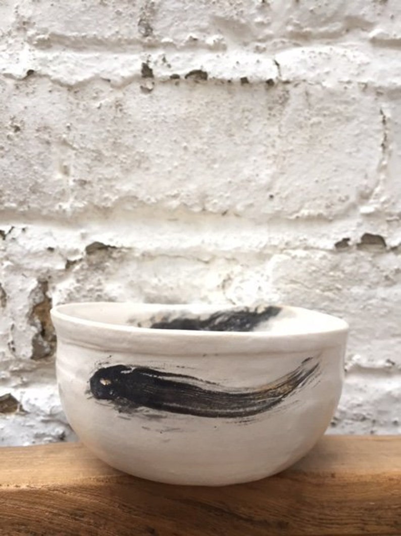 May include: White ceramic bowl with a black and gold brushstroke design. The bowl is sitting on a wooden surface.