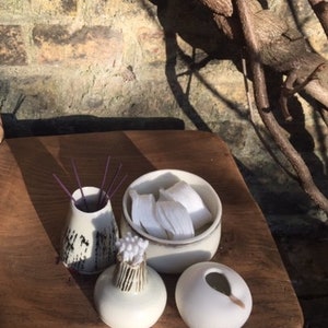 May include: A set of four white ceramic containers with different shapes and designs. The containers are arranged on a wooden surface. One container has a lid and contains white cotton balls. Another container has a black and white pattern and is filled with purple incense sticks. The other two containers are plain white.