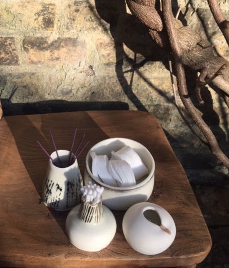 May include: A set of four white ceramic containers with different shapes and designs. The containers are arranged on a wooden surface. One container has a lid and contains white cotton balls. Another container has a black and white pattern and is filled with purple incense sticks. The other two containers are plain white.