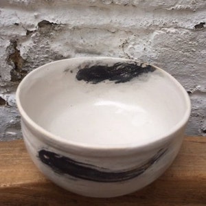 May include: A white ceramic bowl with black brushstrokes on the exterior. The bowl is round and has a smooth, glossy finish.