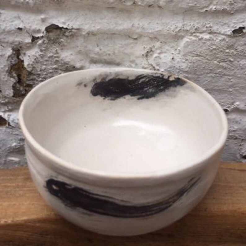 May include: A white ceramic bowl with black brushstrokes on the exterior. The bowl is round and has a smooth, glossy finish.