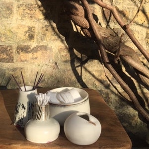 May include: Three white ceramic containers with a rustic, natural look. The containers are different sizes and shapes, with one featuring a black and white pattern. The containers are sitting on a wooden surface.