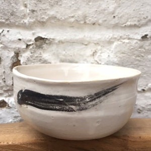 May include: White ceramic bowl with a black brushstroke design. The bowl is round and has a slight lip.