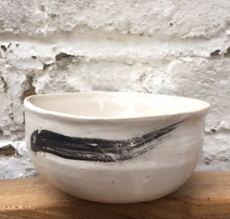 May include: White ceramic bowl with a black brushstroke design. The bowl is round and has a slight lip.