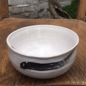 May include: White ceramic bowl with a black brushstroke design. The bowl is round and has a slightly flared rim.