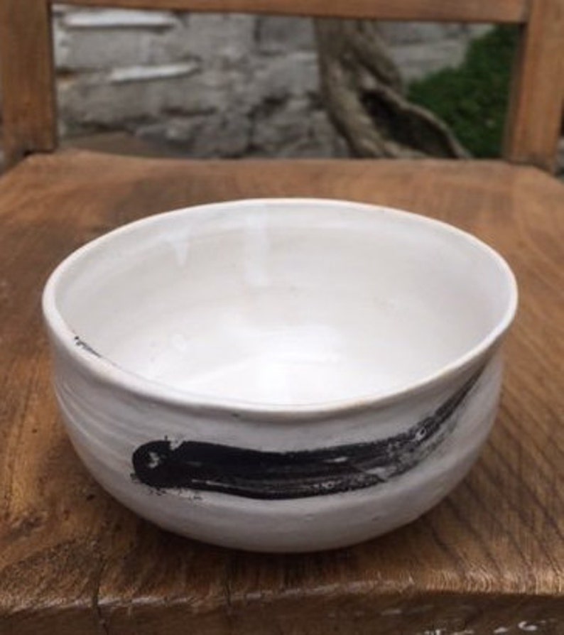 May include: White ceramic bowl with a black brushstroke design. The bowl is round and has a slightly flared rim.