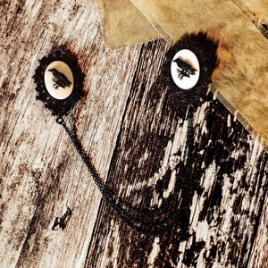 Peut inclure: Deux broches camées noires en forme de corbeau reliées par une chaîne noire. Les broches sont sur une surface en bois rustique avec un tissu brun en arrière-plan.