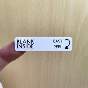 May include: A white rectangular label with black text reading "BLANK INSIDE" on the left and "EASY PEEL" with a curved arrow on the right. The label is held by a finger against a light wood background.