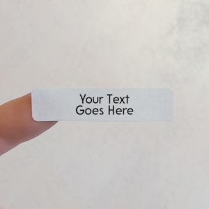 以下が含まれることがあります： 丸みを帯びた角を持つ、小さな長方形の白いラベル。「Your Text Goes Here」というテキストが黒色で印刷され、ラベルの中央に配置されています。ラベルは、無地のオフホワイトの背景に対して指で保持されています。