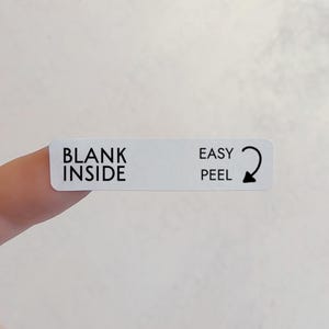 May include: A white rectangular label with black text. The text reads "BLANK INSIDE" on the left and "EASY PEEL" with a curved arrow on the right. The label is held by a finger.