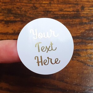 May include: A white, round sticker with gold lettering that reads "Your Text Here." The sticker is held by a finger against a dark wood grain background. The text is in a cursive font.