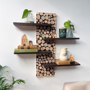 May include: A wooden wall shelf with a unique design featuring a vertical stack of wood rounds. The shelf has four brown shelves with various items on them, including a green candle, a glass vase with a plant, a pitcher, and a stack of books.