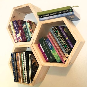 May include: Three hexagonal wooden shelves with books and journals. The shelves are stacked in a honeycomb pattern. The books include titles like "The Witches' Almanac" and "Wiccapedia".