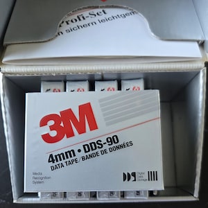 Puede incluir: Una caja plateada que contiene varias cintas de datos 3M 4mm DDS-90. La etiqueta frontal presenta el logotipo rojo de 3M, el texto 4mm DDS-90 DATA TAPE/BANDE DE DONNÉES, el logotipo Media Recognition System y el logotipo Digital Data Storage en blanco y negro.