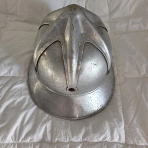 以下が含まれることがあります： クラシックなスタイルの銀色の金属製ヘルメット。頭頂部からつばにかけて伸びる目立つ隆起したクレストデザインが特徴です。ヘルメットは滑らかで丸みを帯びたドーム型で、幅広で広がったつばと、使い込まれたような金属仕上げが施されています。