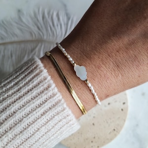 May include: Two bracelets on a wrist. One is a gold chain, the other is a pearl bracelet with a white cloud-shaped charm. The person is wearing a cream-coloured jumper.