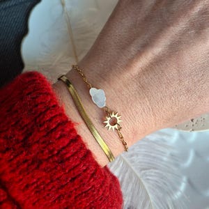 Op de afbeelding: Een gouden armbandenset om een pols. Eén armband is een eenvoudige ketting, de andere heeft een wolkbedel en een zonnebedel. De persoon draagt een rode trui.