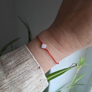 May include: A red string bracelet featuring a small, white, four-petal flower charm. The bracelet is worn on a wrist, with a neutral-toned sleeve in the foreground. The background is blurred, with suggestions of foliage.