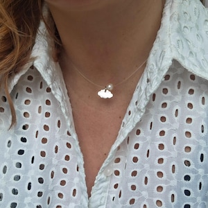 Puede incluir: Un delicado collar de plata en forma de nube con un pequeño dije de perla colgando. El collar lo lleva una persona con una camisa blanca con un patrón de encaje.