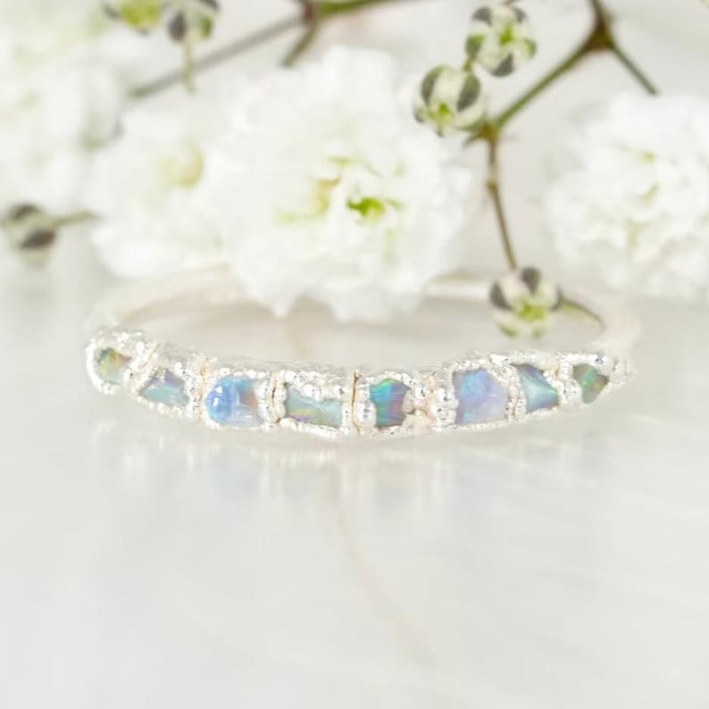Op de afbeelding: Een zilveren ring met een rij iriserende opaalstenen. De opalen hebben blauwe, groene en paarse tinten. De ring is geplaatst tegen een achtergrond van witte bloemen en een wit oppervlak.