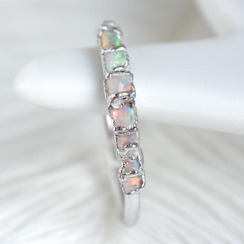 Op de afbeelding: Een zilveren ring met een rij rechthoekige opaalstenen. De opalen vertonen een kleurenspel met flitsen van groen, oranje en roze. De ring is geplaatst tegen een zachte, witte achtergrond, die de edelstenen benadrukt.