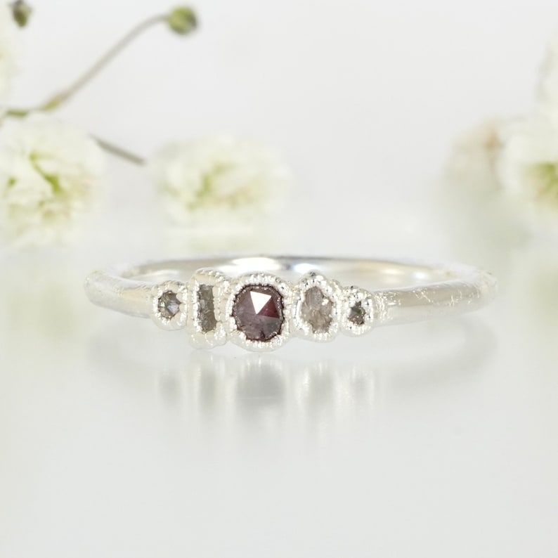 May include: A silver band ring featuring five small, irregularly shaped diamonds. The central diamond is a dark grey colour, while the other four diamonds are a lighter grey colour.