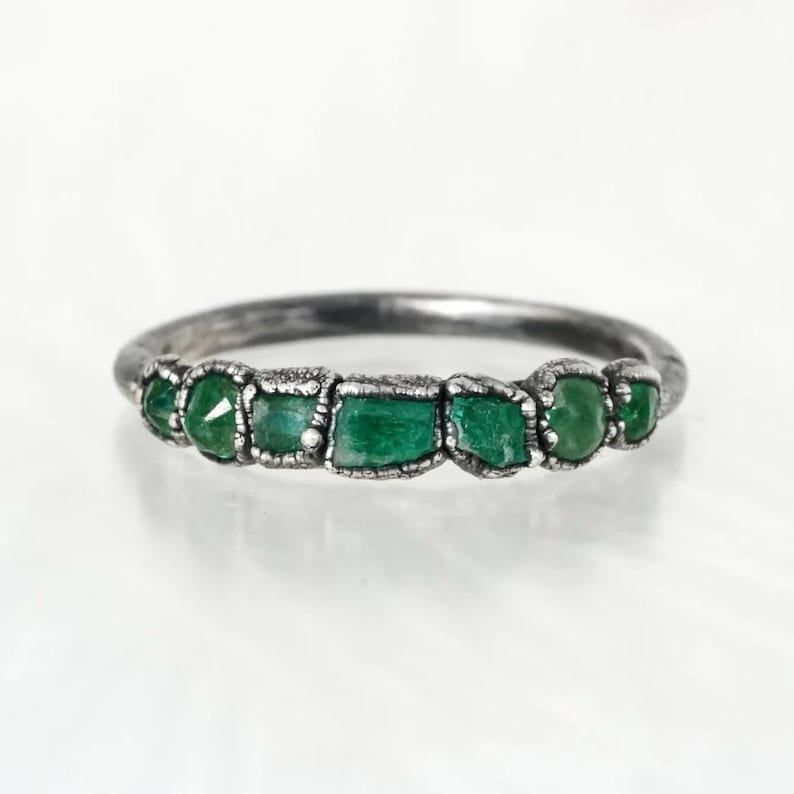 May include: A silver ring with a row of six green gemstones set in the band. The gemstones are irregularly shaped and have a rough, natural look.