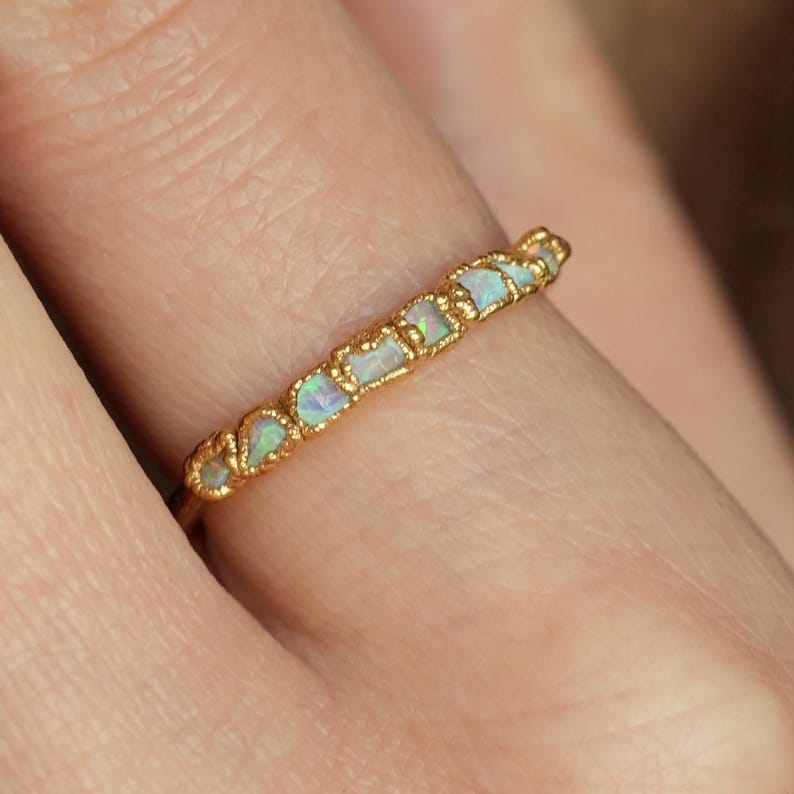 Op de afbeelding: Een gouden ring met een rij kleine, onregelmatige opaalstenen. De opalen vertonen lichtblauwe, groene en witte tinten. De band van de ring heeft een textuurrijk, sierlijk ontwerp.