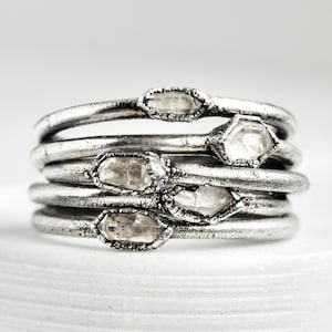 May include: A collection of silver rings, each featuring a raw, clear crystal. The rings have a textured, oxidised finish, and the crystals are set in a bezel. The rings are stacked on a white surface.