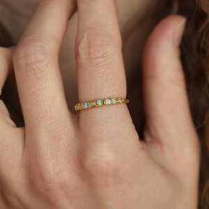 Op de afbeelding: Een gouden ring met een reeks kleine, rechthoekige opalen. De opalen vertonen een subtiele iriserende kwaliteit, met hints van blauw en groen. De ring wordt aan een vinger gedragen.