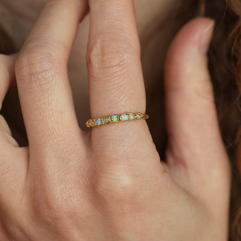 Op de afbeelding: Een gouden ring met een reeks kleine, rechthoekige opalen. De opalen vertonen een subtiele iriserende kwaliteit, met hints van blauw en groen. De ring wordt aan een vinger gedragen.