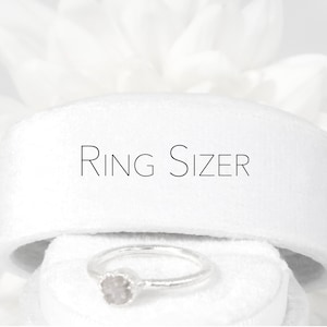 Puede incluir: Un medidor de talla de anillo blanco con el texto "RING SIZER" impreso en él. Un anillo de plata con una piedra transparente está sentado encima del medidor.