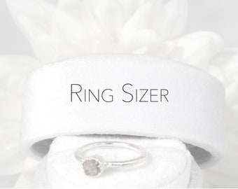 May include: A white ring sizer with the text "RING SIZER" printed on it. A silver ring with a clear stone is sitting on top of the sizer.