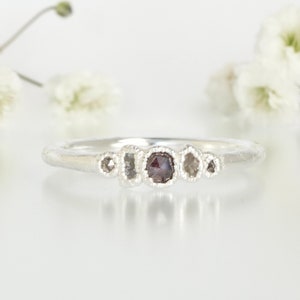 May include: A silver ring featuring five small, irregularly shaped diamonds. The central diamond is a dark grey colour, while the other four diamonds are clear.