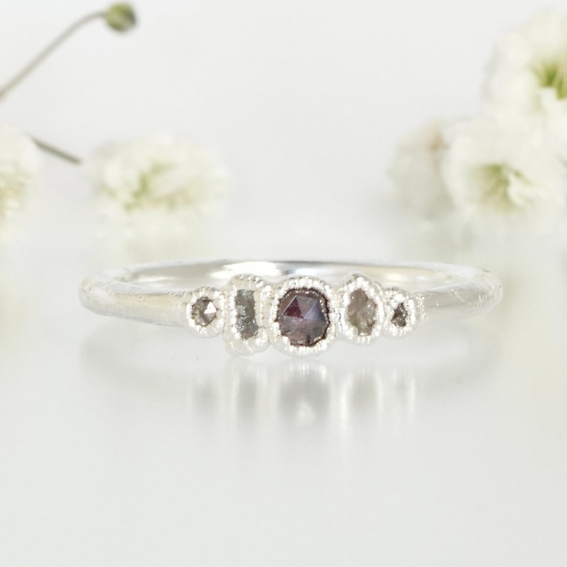 May include: A silver ring featuring five small, irregularly shaped diamonds. The central diamond is a dark grey colour, while the other four diamonds are clear.