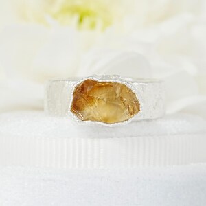 May include: A silver ring featuring a raw, golden-yellow citrine gemstone. The ring has a textured band and a bezel setting that highlights the natural shape of the stone. The citrine has a rough, organic appearance.